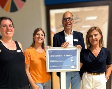 Vier Personen stehen zusammen und halten ein Schild mit der Aufschrift "Herzlich Willkommen" in der Hand.
