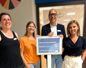 Vier Personen stehen zusammen und halten ein Schild mit der Aufschrift "Herzlich Willkommen" in der Hand.