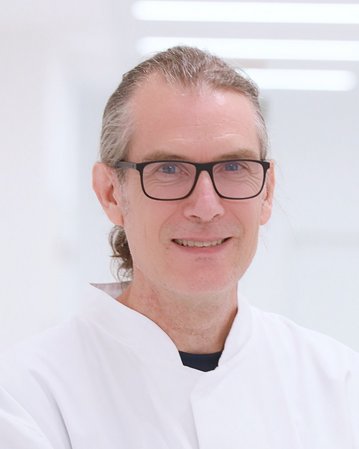 Ein Mann mit Brille und langem, grauem Haar trägt einen weißen Kittel. Er lächelt freundlich in einem hellen, modernen Raum.