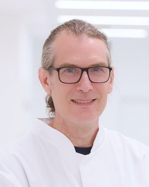 Ein Mann mit Brille und langem, grauem Haar trägt einen weißen Kittel. Er lächelt freundlich in einem hellen, modernen Raum.