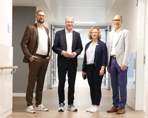Vier Personen stehen in einem modernen Flur, gekleidet in formelle und legere Kleidung, und schauen in die Kamera.