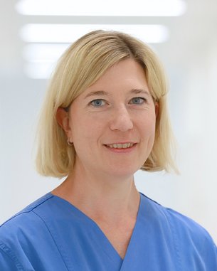 Eine Frau mit blonden Haaren trägt einen blauen Kittel und lächelt freundlich. Der Hintergrund ist hell und unscharf.