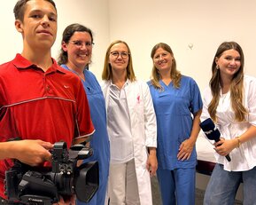 Eine Gruppe von fünf Personen, darunter ein Mann mit einer Kamera, steht in einem Raum. Zwei Frauen tragen medizinische Kleidung, während die anderen lässige Kleidung tragen.