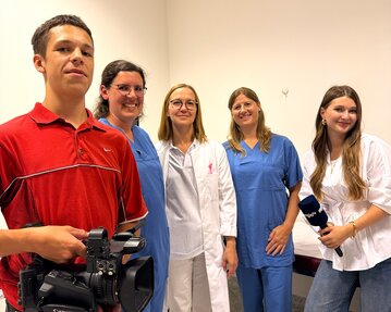 Eine Gruppe von fünf Personen, darunter ein Mann mit einer Kamera, steht in einem Raum. Zwei Frauen tragen medizinische Kleidung, während die anderen lässige Kleidung tragen.