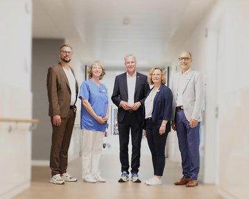 Fünf Personen stehen in einem Krankenhausflur. Zwei tragen medizinische Kleidung, während die anderen formelle Kleidung tragen.