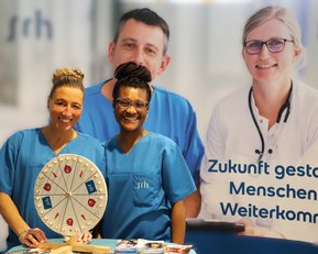 Zwei Frauen in blauen Arbeitskleidung stehen vor einem Banner mit dem Text „Zukunft gestalten, Menschen helfen, Weiterkommen“ und einem Glücksrad.