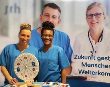 Zwei Frauen in blauen Arbeitskleidung stehen vor einem Banner mit dem Text „Zukunft gestalten, Menschen helfen, Weiterkommen“ und einem Glücksrad.