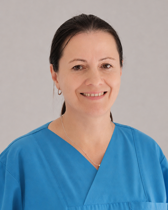 Eine Frau in blauer Arbeitskleidung lächelt freundlich. Sie hat lange, dunkle Haare und trägt Ohrringe.