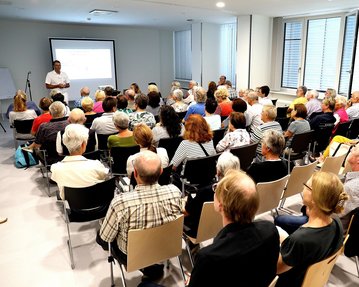 Eine große Gruppe von Menschen sitzt in einem Raum und hört einem Vortragenden zu, der vor einer Präsentation steht.