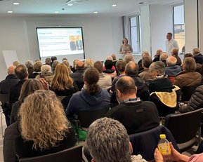 Eine Gruppe von Menschen sitzt in einem Konferenzraum und hört einer Präsentation zu, die auf einem Bildschirm gezeigt wird.