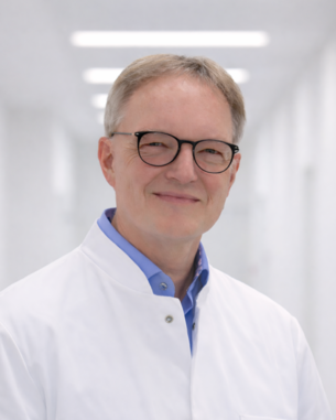 Ein lächelnder Arzt in einem weißen Kittel steht in einem hellen Flur eines Krankenhauses.