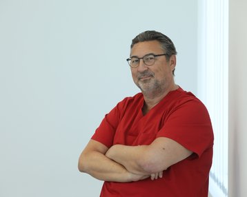 Ein Mann in roter Arztkleidung steht mit verschränkten Armen und lächelt. Er trägt eine Brille und hat graue Haare.