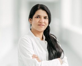 Eine Frau in einem weißen Kittel steht mit verschränkten Armen in einem hellen Flur. Sie hat lange, schwarze Haare und schaut direkt in die Kamera.