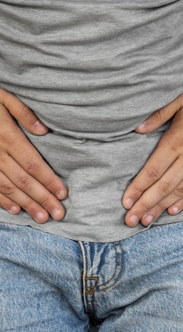 Mann mit grauem T-Shirt hält sich den Bauch, möglicherweise aufgrund von Unwohlsein oder Schmerzen.