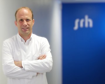 Ein Arzt in einem weißen Kittel steht mit verschränkten Armen vor einer blauen Wand mit dem Schriftzug "srh".