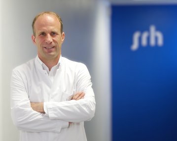 Ein Arzt in einem weißen Kittel steht mit verschränkten Armen vor einer blauen Wand mit dem Schriftzug "srh".