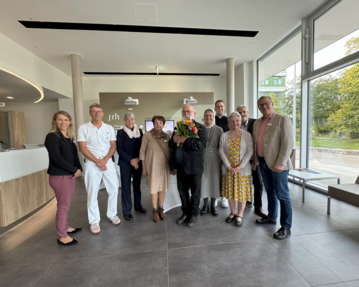 Eine Gruppe von zehn Personen steht in einem modernen Empfangsbereich, einige halten Blumen. Sie scheinen eine Feier oder ein besonderes Ereignis zu haben.