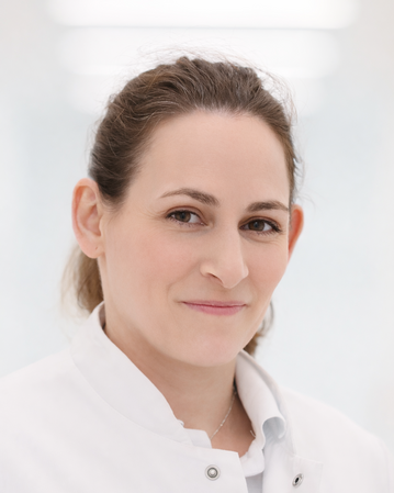 Eine Frau in einem weißen Kittel lächelt freundlich. Ihr Haar ist leicht gewellt und sie trägt eine Halskette.