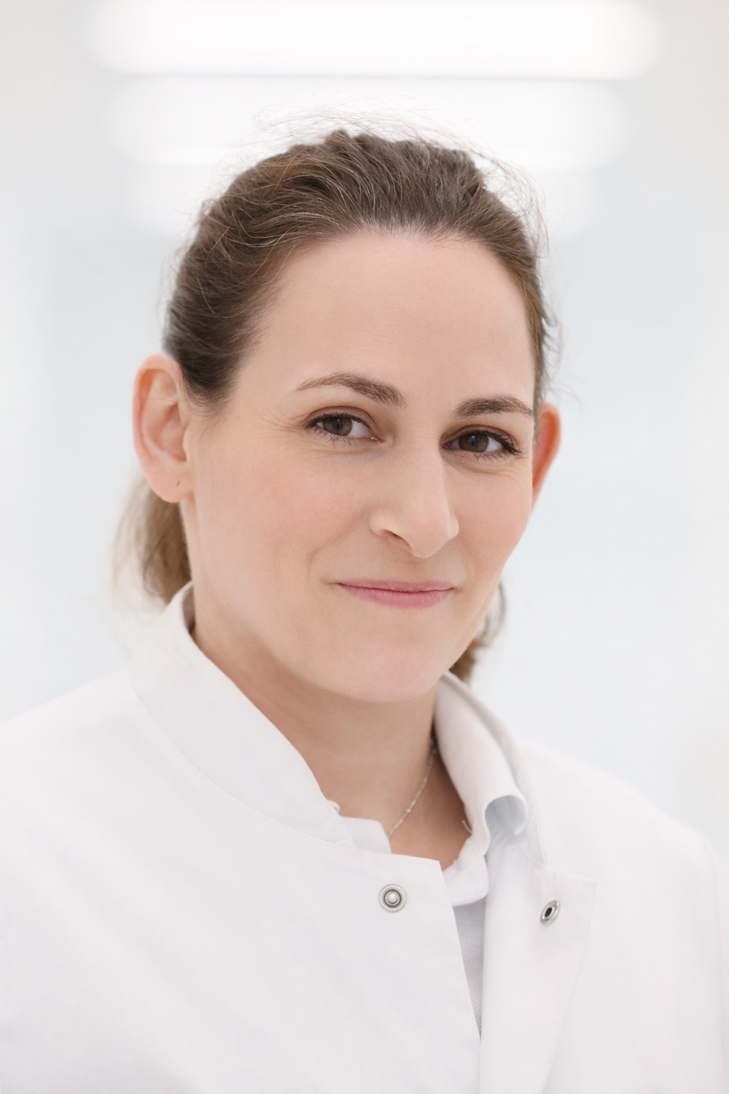 Eine Frau in einem weißen Kittel lächelt freundlich. Ihr Haar ist leicht gewellt und sie trägt eine Halskette.