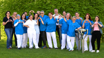 Eine Gruppe von 15 Personen in blauen T-Shirts formt mit ihren Händen Herzen. Sie stehen im Freien vor einer grünen Wand aus Pflanzen.