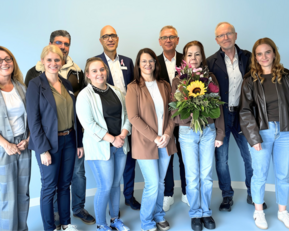 Eine Gruppe von zehn Personen steht zusammen, einige halten Blumen. Der Hintergrund ist hellblau.