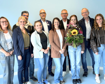 Eine Gruppe von zehn Personen steht zusammen, einige halten Blumen. Der Hintergrund ist hellblau.