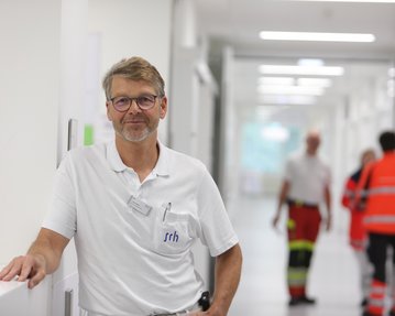 Ein Arzt in einem weißen Kittel steht in einem modernen Krankenhausflur, während im Hintergrund Rettungskräfte vorbeigehen.
