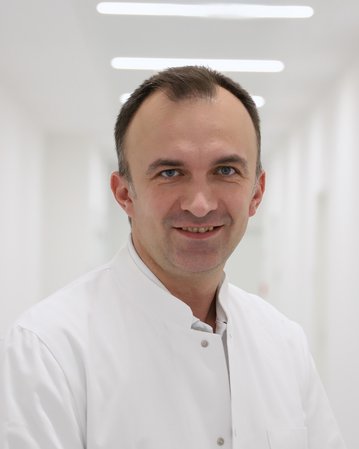 Ein Arzt in einem weißen Kittel lächelt freundlich in einem hellen Flur eines Krankenhauses.