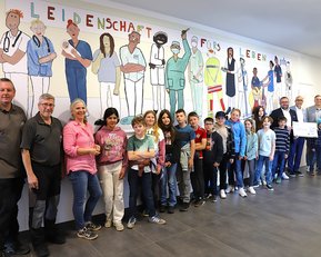 Eine Gruppe von Erwachsenen und Kindern steht vor einem Wandgemälde, das verschiedene Berufe darstellt, darunter Ärzte und Pflegekräfte.