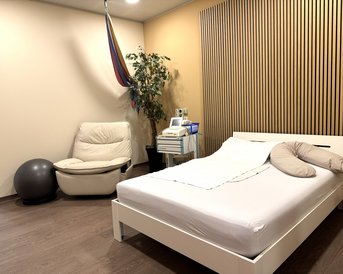 Ein Schlafzimmer mit einem weißen Bett, einem Sessel, einem Gymnastikball und einer Pflanze. Eine Hängematte ist an der Wand befestigt.