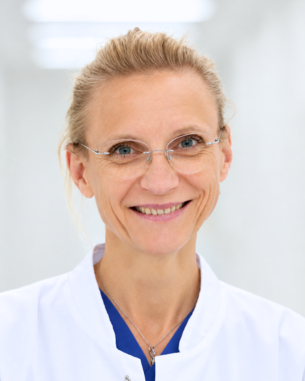 Eine lächelnde Ärztin trägt eine weiße Kittel und eine Brille, steht vor einem hellen, neutralen Hintergrund.