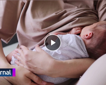 Eine Mutter hält ein schlafendes Baby in ihren Armen, während sie in einem hellen Raum sitzt. Ein Logo mit der Aufschrift "journal" ist in der Ecke sichtbar.