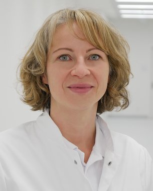 Eine Frau mit blonden, lockigen Haaren trägt einen weißen Kittel. Sie steht in einem hellen, modernen Flur mit neutralen Wänden. Ihr Blick ist freundlich und direkt in die Kamera gerichtet.