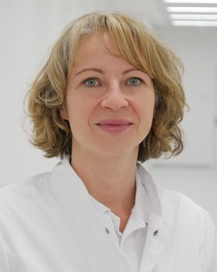 Eine Frau mit blonden, lockigen Haaren trägt einen weißen Kittel. Sie steht in einem hellen, modernen Flur mit neutralen Wänden. Ihr Blick ist freundlich und direkt in die Kamera gerichtet.