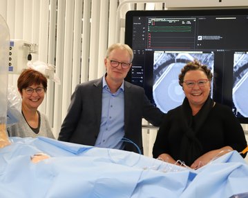 Drei Personen stehen lächelnd vor einem medizinischen Bildschirm, der medizinische Bilder zeigt. Sie tragen Krankenhauskleidung und sind in einem Behandlungsraum.