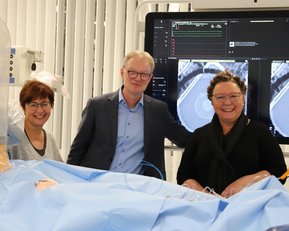 Drei Personen stehen lächelnd vor einem medizinischen Bildschirm, der medizinische Bilder zeigt. Sie tragen Krankenhauskleidung und sind in einem Behandlungsraum.