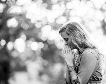 Eine Frau mit langen Haaren steht mit gefalteten Händen und gesenktem Kopf, umgeben von unscharfen, hellen Lichtern im Hintergrund.