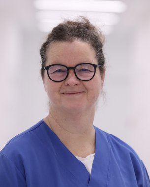 Eine Frau mit Brille trägt einen blauen Kittel und lächelt. Der Hintergrund ist hell und unscharf, was den Fokus auf die Person legt.