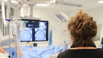Eine Person mit lockigem Haar steht vor einem medizinischen Bildschirm, der zwei runde Bilder zeigt, während ein medizinisches Team im Hintergrund arbeitet.