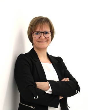 Eine lächelnde Frau mit Brille steht mit verschränkten Armen vor einer weißen Wand, trägt einen schwarzen Blazer und ein weißes Oberteil.