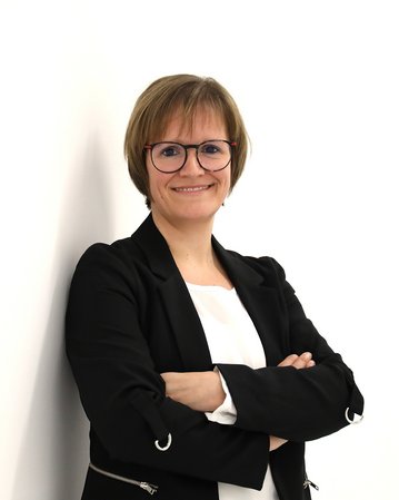 Eine lächelnde Frau mit Brille steht mit verschränkten Armen vor einer weißen Wand, trägt einen schwarzen Blazer und ein weißes Oberteil.