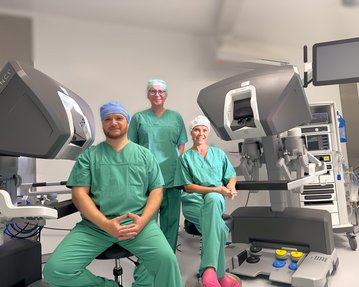 Drei medizinische Fachkräfte in grüner OP-Kleidung sitzen und stehen neben einem chirurgischen Robotersystem in einem Operationssaal.