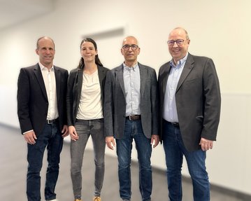 Vier Personen stehen in einem hellen Raum, lächelnd und in formeller Kleidung, bereit für ein Treffen oder eine Veranstaltung.