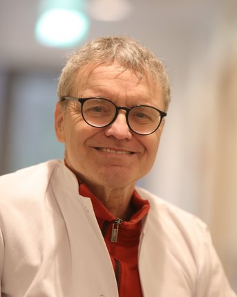 Ein lächelnder Arzt in einem weißen Kittel, mit Brille und rotem Oberteil, sitzt in einem hellen Raum.