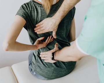 Eine Person sitzt auf einer Liege, während eine andere Person mit beiden Händen den Rücken der sitzenden Person behandelt.