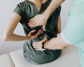 Eine Person sitzt auf einer Liege, während eine andere Person mit beiden Händen den Rücken der sitzenden Person behandelt.