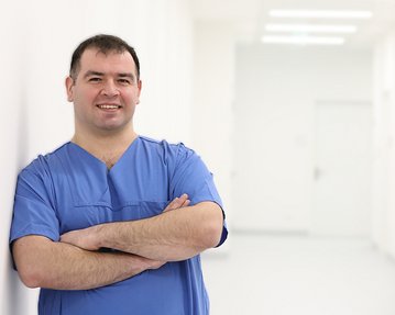 Ein männlicher Arzt in blauer Arbeitskleidung steht mit verschränkten Armen in einem hellen Flur.