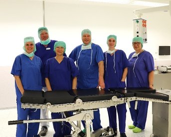 Sechs medizinische Fachkräfte in blauen OP-Kitteln und Hauben stehen um eine chirurgische Liege in einem Operationssaal.