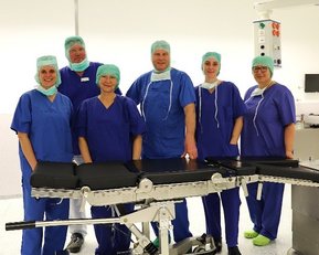 Sechs medizinische Fachkräfte in blauen OP-Kitteln und Hauben stehen um eine chirurgische Liege in einem Operationssaal.