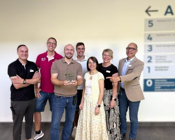Eine Gruppe von sieben Personen steht zusammen, einige halten einen Preis. Im Hintergrund sind nummerierte Schilder sichtbar.